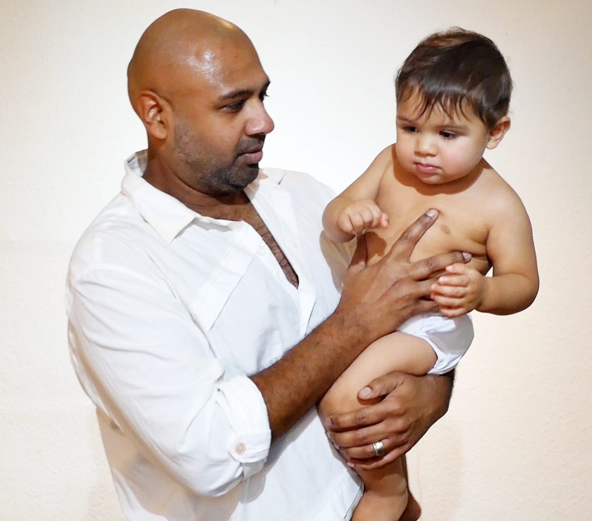 A South Asian man in a white shirt holds his young son in his arms
